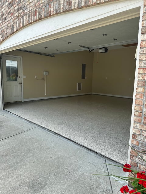 Empty garage interior with open door, tan walls, concrete floor, and red flowers in foreground