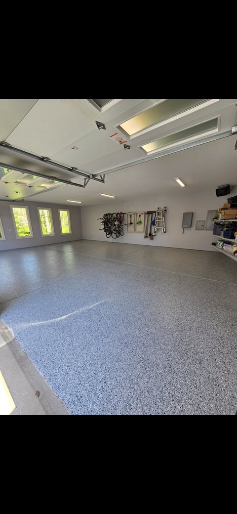 Spacious garage interior with speckled gray epoxy floor, overhead doors with windows, bright LED lighting, wall-mounted bikes and tools, and clear organizational storage on right side.